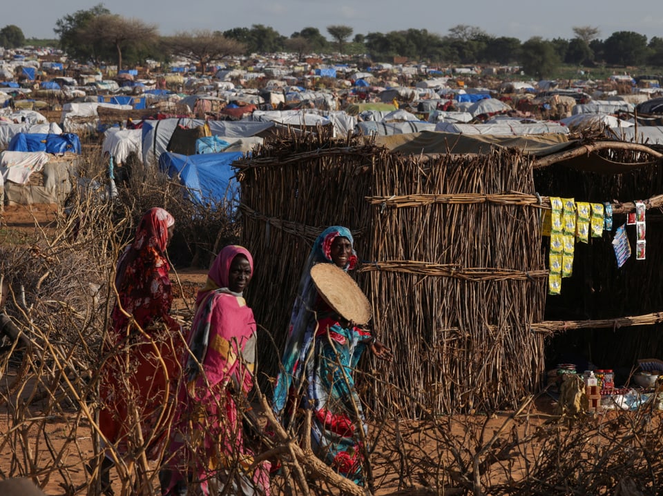 Colorfully dressed women walk past a straw hut in a refugee camp.