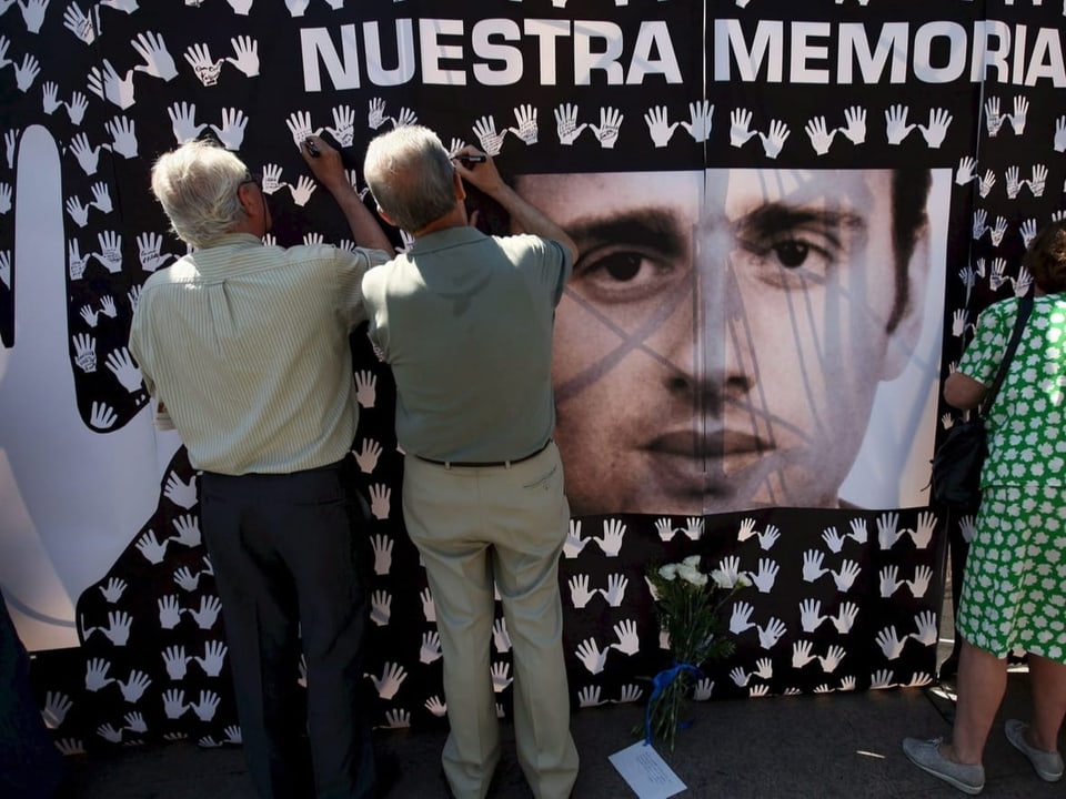 Drei Menschen schreiben draussen Botschaften neben ein grosses Porträt von Miguel Ángel Blanco.