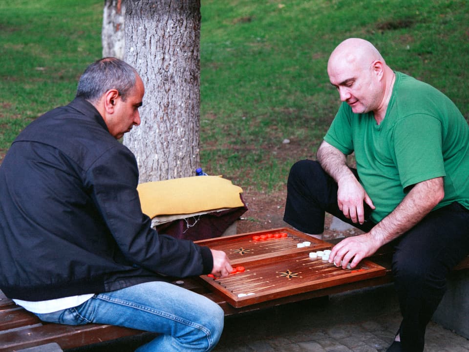 Zwei Männer sitzen auf einer Bank und spielen ein Brettspiel.