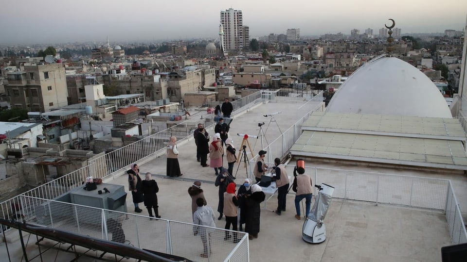 Menschen auf einem Dach mit Teleskopen. Man sieht eine Moschee.