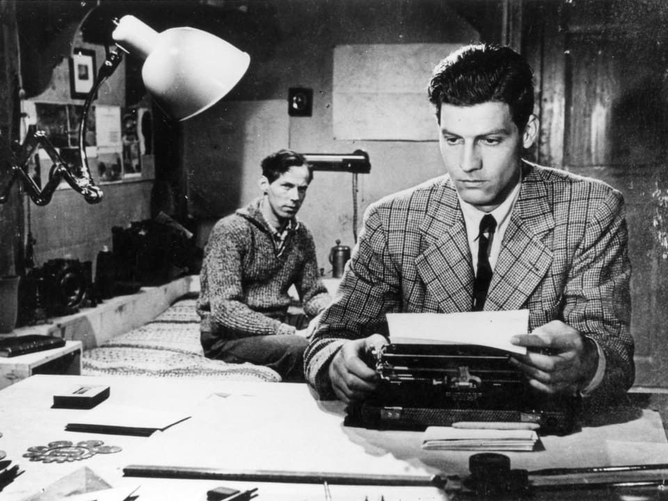 Ein Mann sitzt an einer Schreibmaschine und hält inne. Ein zweiter Mann sitzt hinter ihm im Raum und beobachtet ihn.