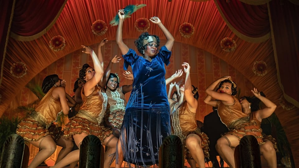 Dunkelhäutige Frauen tanzen auf einer Bühne. In der Mitte steht eine Frau im blauen Samtkleid und mit einem Fächer.
