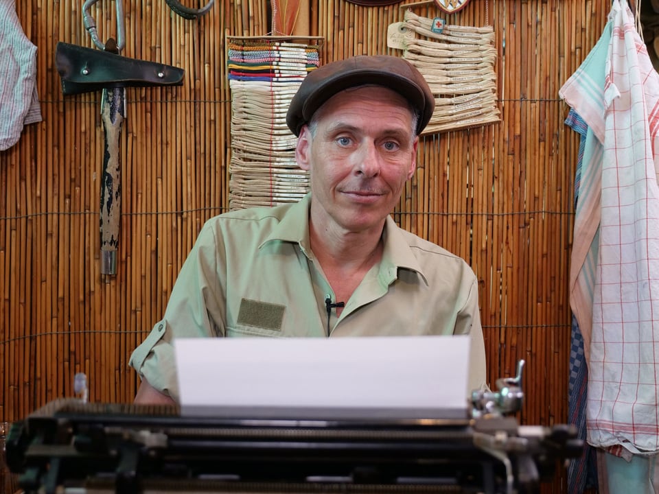 Ein Mann mit Schiebermütze sitzt lächelnd hinter einer alten Schreibmaschine.