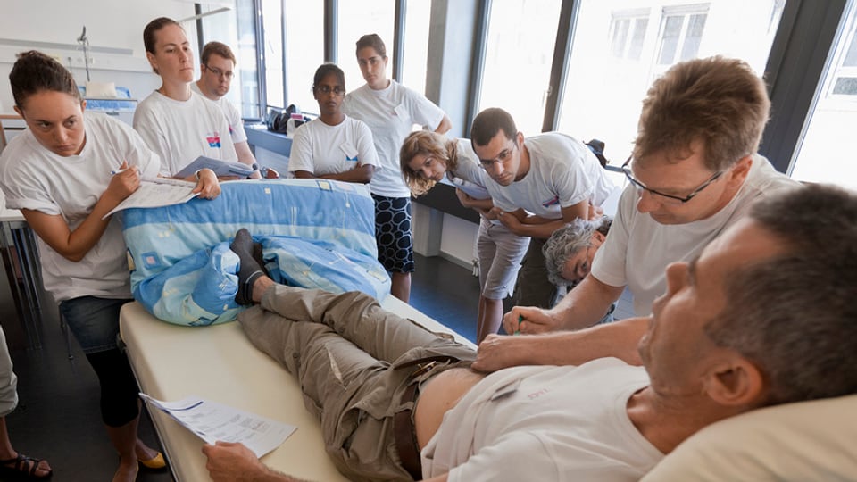 Sieben Auszubildende beobachten an einem Spitalbett den Ausbildner bei der Patientenpflege.