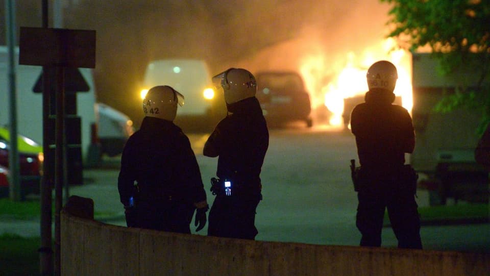 Drei Polizisten mit Helm beobachten ein brennendes Auto aus sicherer Entfernung.