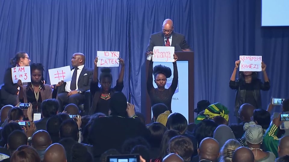 Frauen halten Schilder, Präsident Zuma spricht auf dem Podium