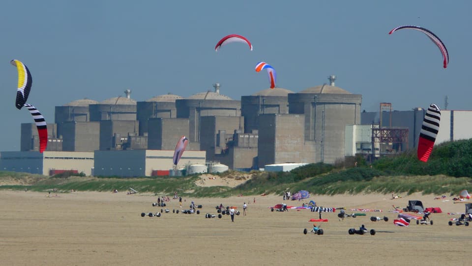 Sechs grosse Betonreaktoren stehen hinter einem Strand, auf dem Personen Drachen steigen lassen.
