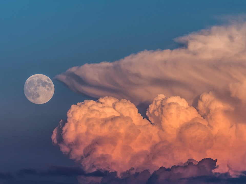 In der rechten Bildhälfte steht ein Gewitterturm im Sonnenuntergang. Links liegt der Mond.