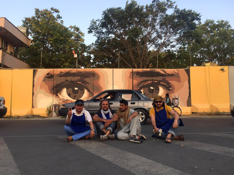 Vier Männer sitzen auf der Strasse vor einem parkiertem Auto. Im Hintergrund ist eine bemalte Mauer erkennbar.
