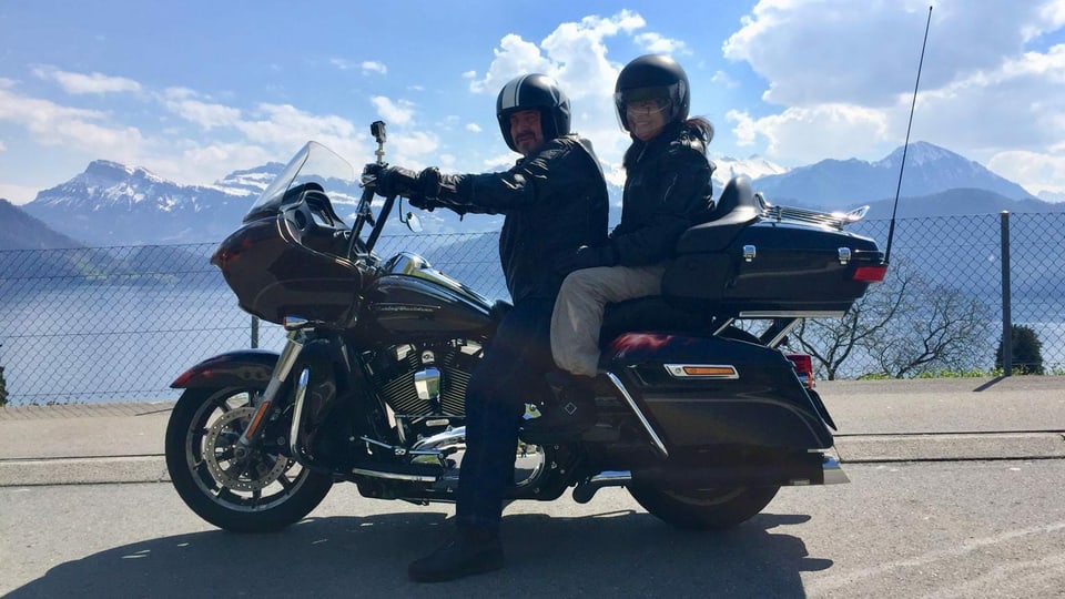 Ein Mann und eine Frau sitzen auf einem grossen Motorrad vor einer See- und Berglandschaft.