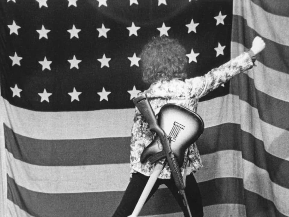 Ein Mann mit Blumenhemd und Gitarre sowie Waffe steht vor einer amerikanischen Flagge und hebt die geballte Faust