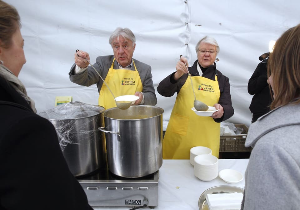 Mann mit weissen Haaren und Frau mit weissen haaren schöpfen in einem Zelt Suppe, haben gelbe Schürzen an.