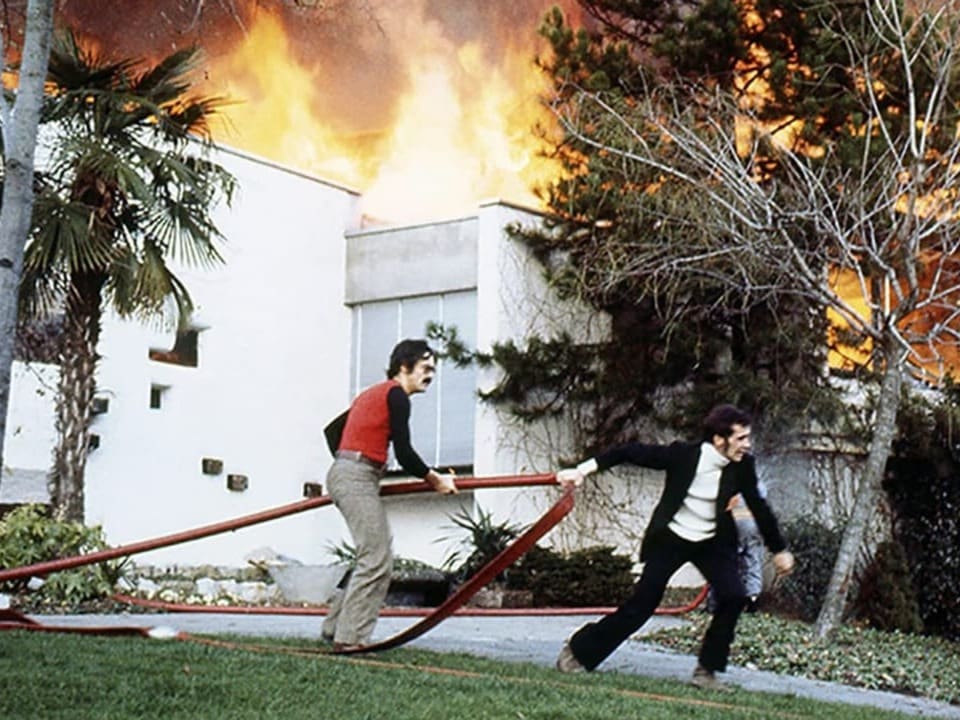 Zwei Männer mit Feuerwehrschlauch vor brennendem Gebäude.