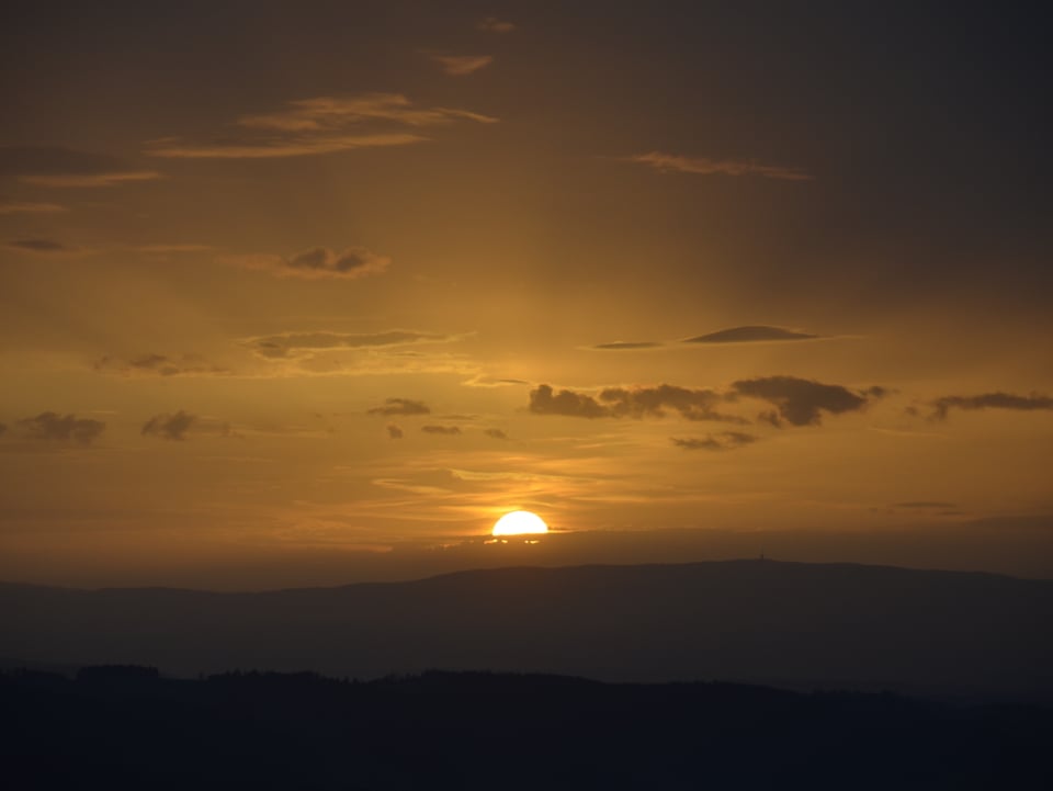 Im unteren Bilddrittel ist die zarte Hügellandschaft des Bernbiets im Schatten zu sehen. Am Horizont ist die Sonne halb untergegangen, und erleuchtet in sanftem Licht den Abendhimmel.