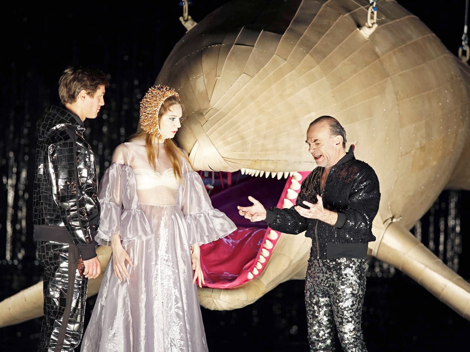 Zwei Männer und eine Frau auf der Theaterbühne, neben ihnen ein Fisch aus Holz.