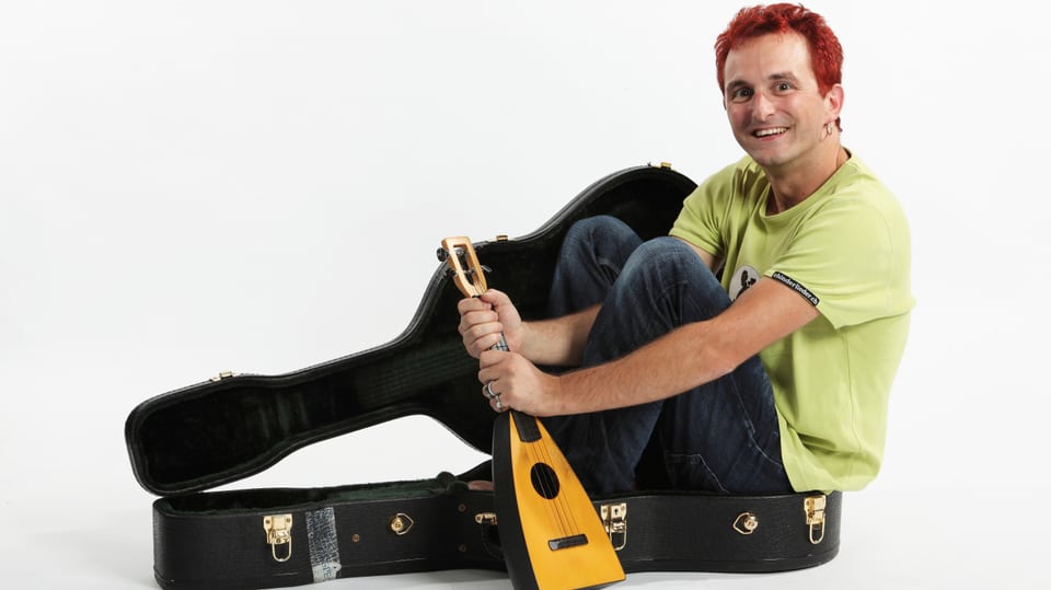 Christian Schenke mit roten Haaren sitzt in einem Gitarrenkoffer und hällt eine Ukulele.