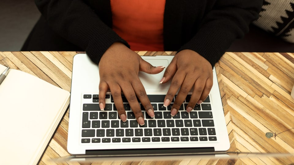 Person tippt auf einem Laptop mit sichtbaren Händen und einem Notizbuch daneben.