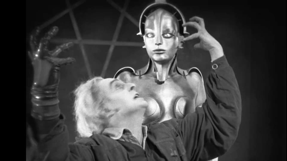 Ein metallischer Roboter mit Frauengesicht steht hinter einem Mann, der exstatisch die Hände verwirft.
