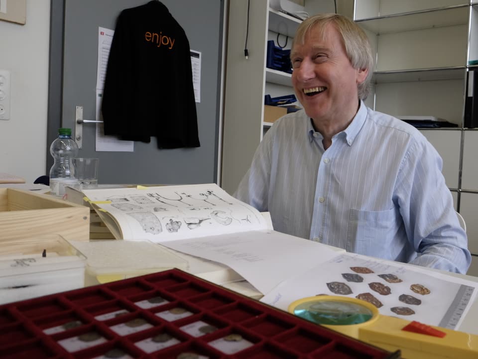 Der Numismatiker Stephen Doswald sitzt lachend am Tisch mit dem Münzfund und verschiedenen Büchern.