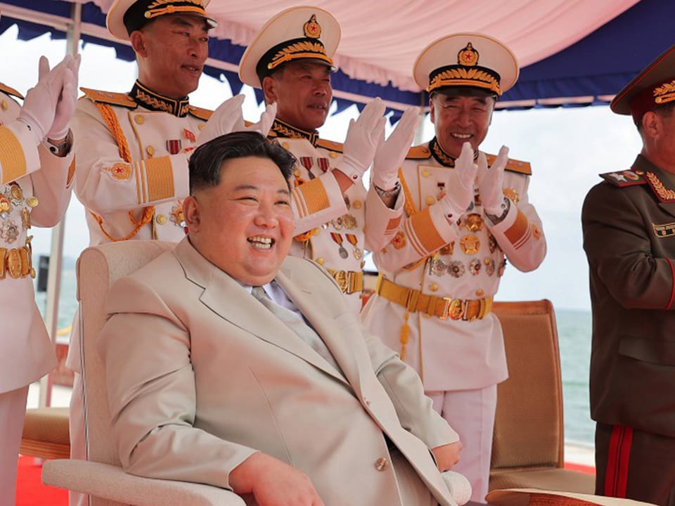 Kim Jong-un sitzt auf einem Stuhl, hinter ihm stehen klatschende Soldaten. 