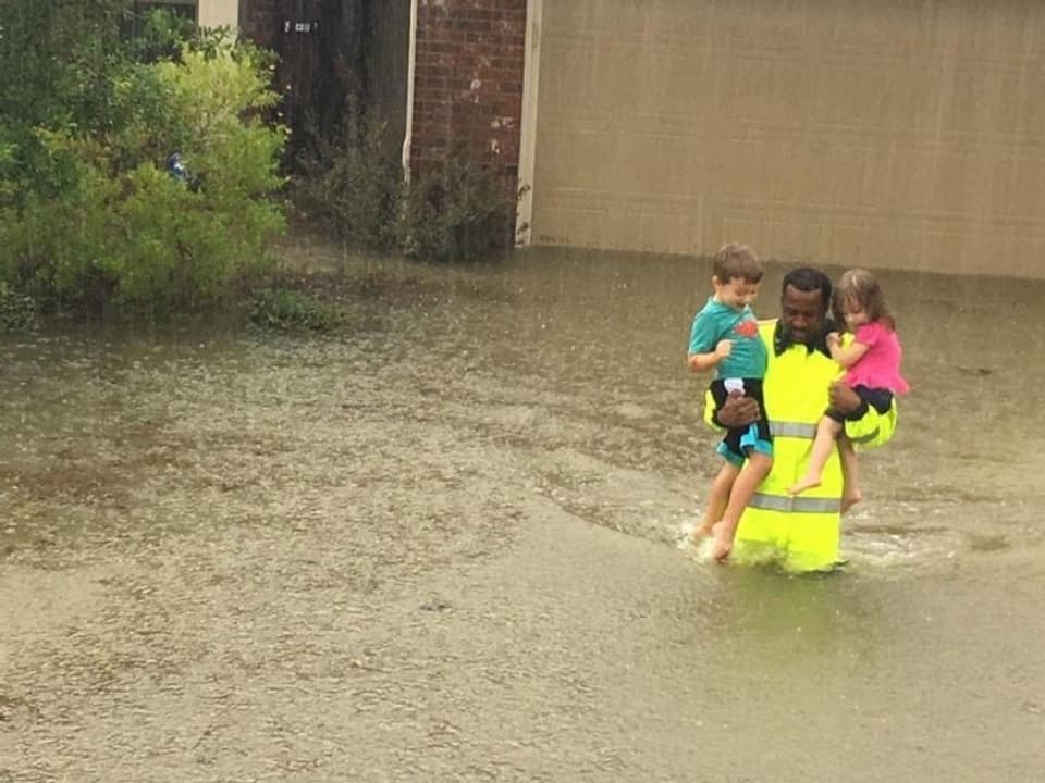 Ein Polizist mit neongelber Weste trägt zwei Kinder auf den Armen und geht durch hüfthohes Wasser.