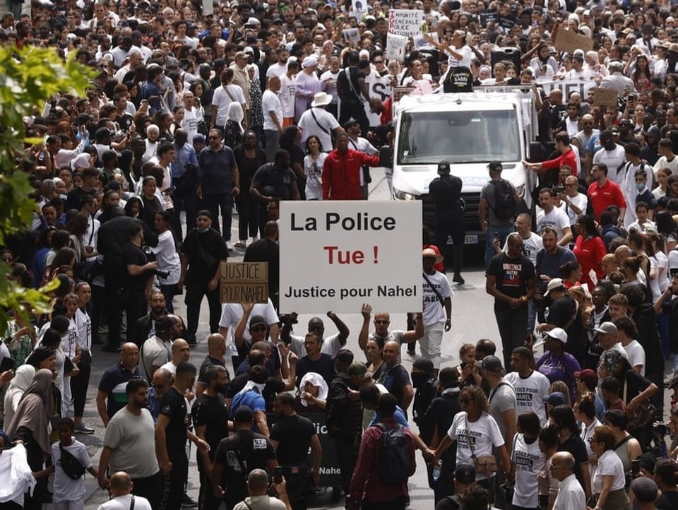 Viele Menschen Demonstrieren. Zwei Menschen halten ein Plakat mit der Aufschrift: «La Police Tue! Justice pour Nahel»