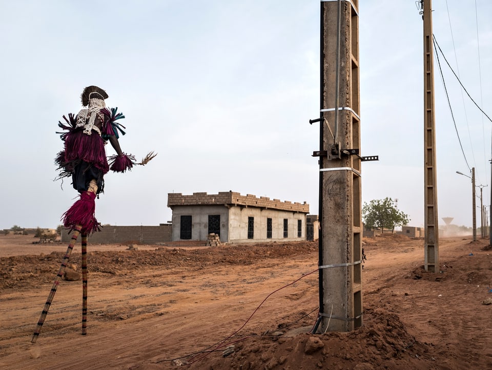 Strassenszene aus Mali mit einem vogelartig verkleideten Menschen, der auf Stelzen geht.