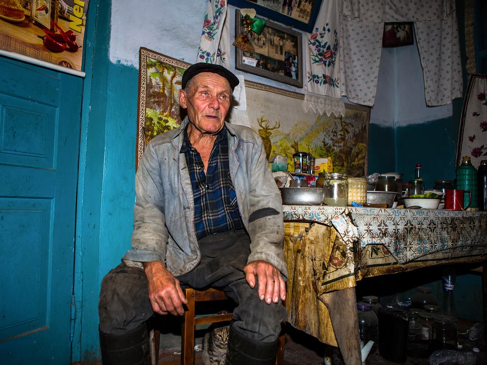 Ein älterer Mann in schmutzigen Arbeitskleidern sitzt an einem überfüllten Küchentisch.