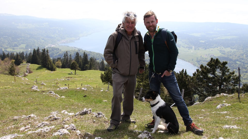 Jean-Louis Raymond und Nik Hartmann vor Aussicht auf den Lac de Joux mit Hund zu Füssen. 