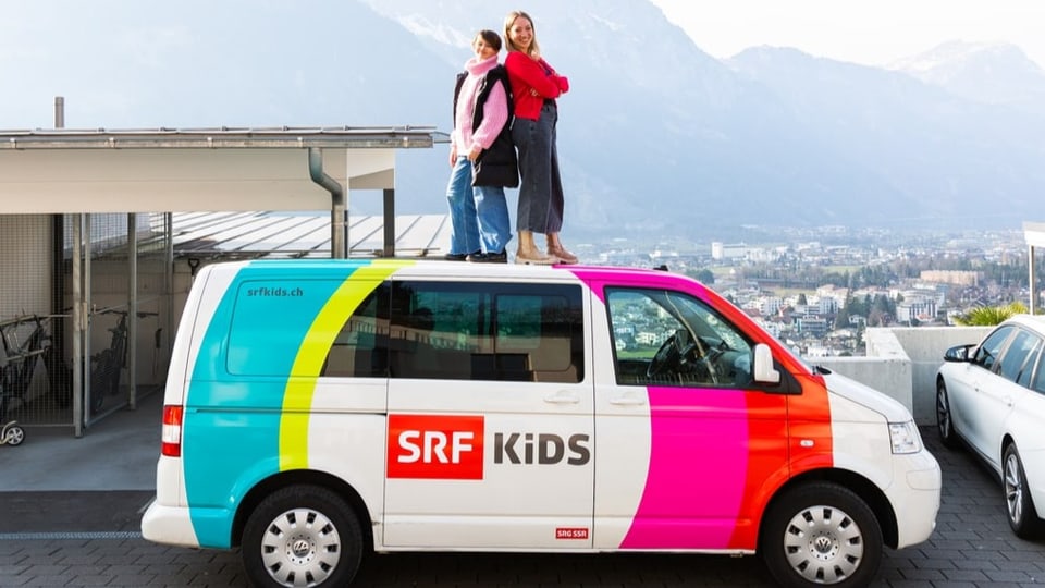 Kommt mit SRF Kids auf Reportage! Kinderreporterin Anouk und SRF Kids Moderatorin Anna Zöllig stehen auf dem Dach des SRF Kids Bus