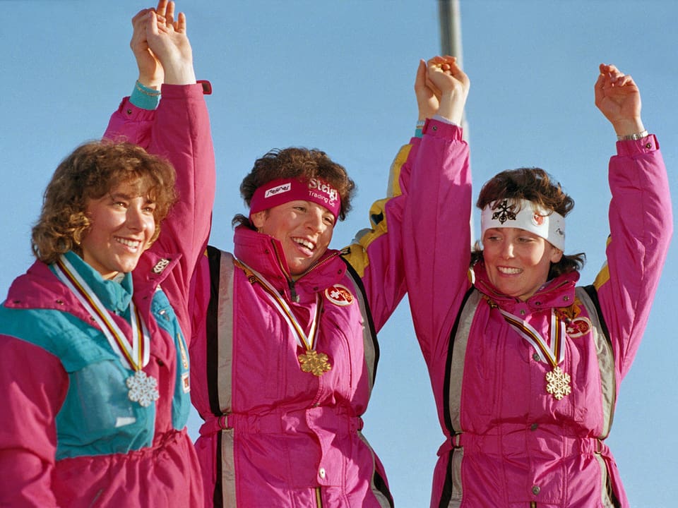 Drei Frauen stehen in pinken Skidresses auf dem Podest und halten die Arme in die Höhe.