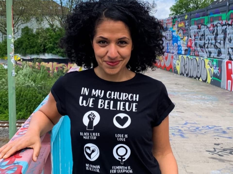 Junge Frau mit dunklen Locken und schwarzem T-Shirt  auf Asphalt-Brücke, Hand auf dem hellblauen Geländer, lächelt.