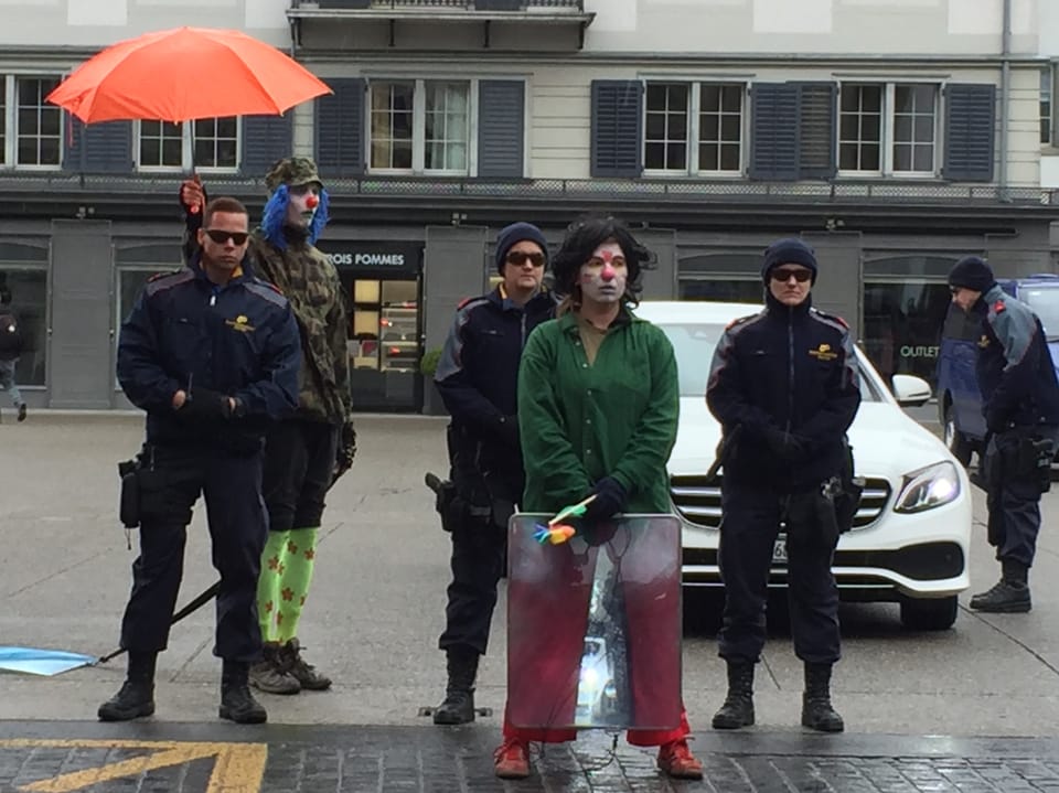 Ein Clown ahmt die Haltung der Polizisten nach, ein zweiter Clown schützt einen Polizisten mit einem orangen Schirm.