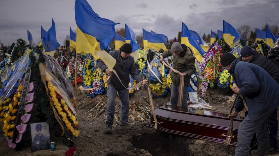 Auf einem Friedhof wird ein Sarg in die Erde gesenkt, Dutzende ukrainische Flaggen wehen auf dem ganzen Friedhof.