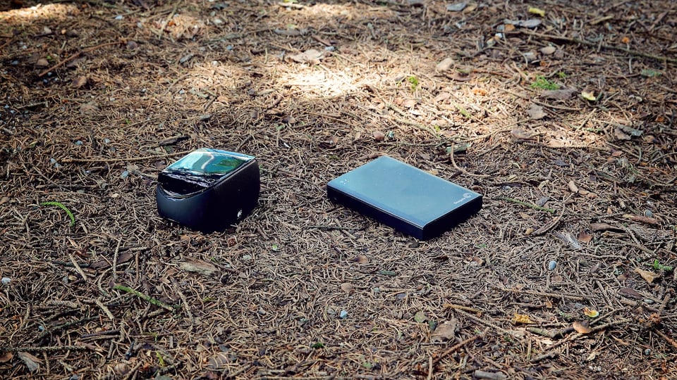 Ein Bluetooth-Lautsprecher und eine Akku-Festplatte liegen am Waldboden.