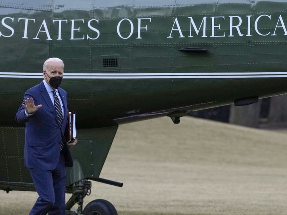 Joe Biden steht vor einem Flugzeug und hebt eine Hand. Er trägt eine Hygienemaske.