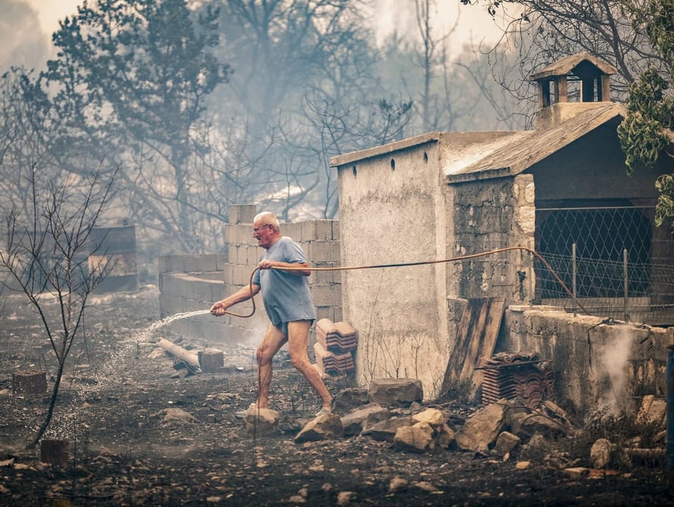 A man sprays a water hose on a burning ground.  He is not wearing proper pants.