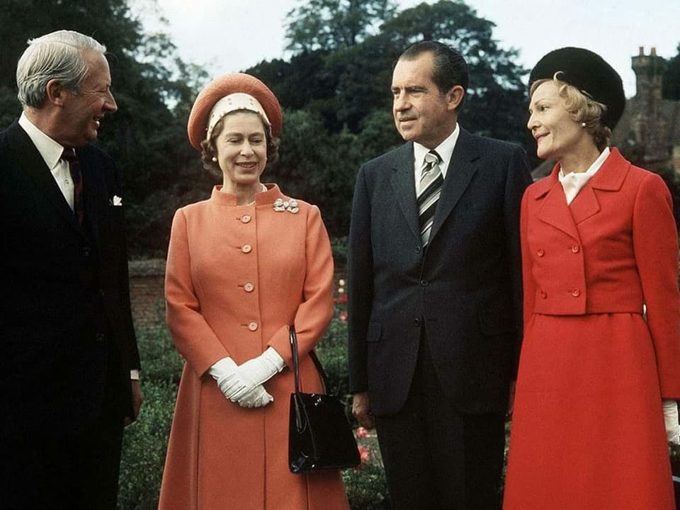 Vier Menschen stehen im Garten zusammen: Die beiden Frauen tragen rote Mäntel und Röcke, die Männer schwarze Anzüge. Die Stimmung ist freundlich.