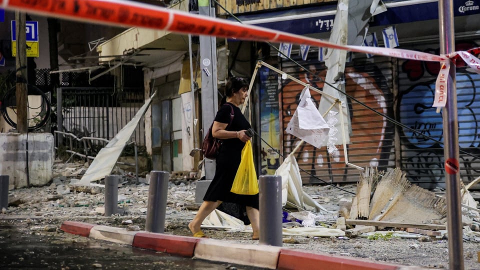 Eine Frau läuft mit gelber Plastiktüte hinter Absperrbändern über einen Strassenabschnitt mit zerstörten Gebäuden.