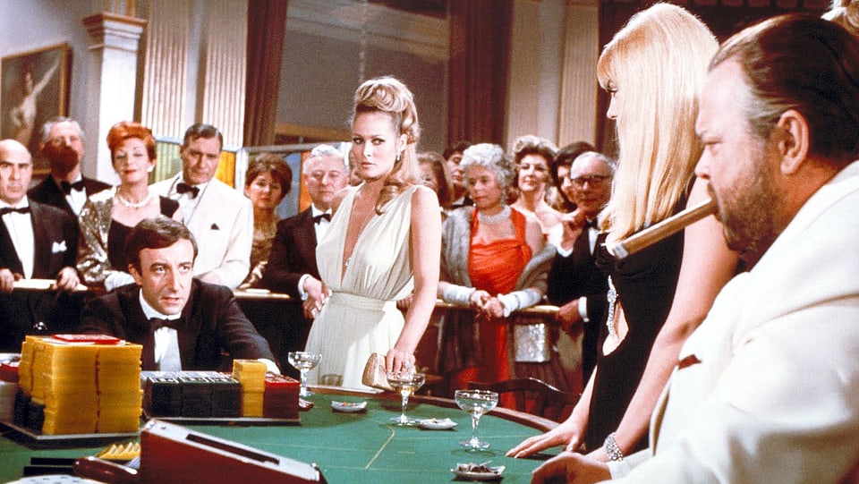 Ursula Andress, Orson Welles, Peter Sellers an einem Pokertisch.