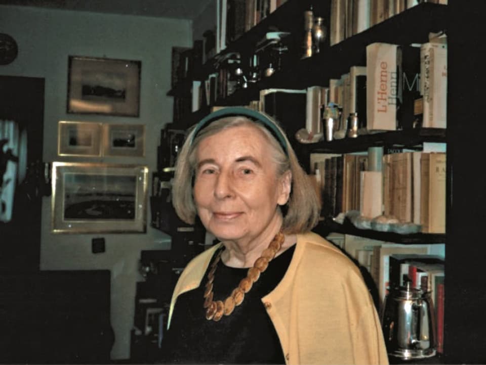 Porrätbild einer älteren Frau mit Haarreif und gelber Strickjacke. Im Hintergrund: Eine Bücherwand und Bilder
