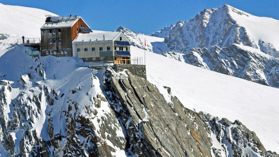 Bild von der Hollandiahütte im Schnee. Im Hintergrund ein Berg. Von der Hütte fällt ein Felsen steil ab.