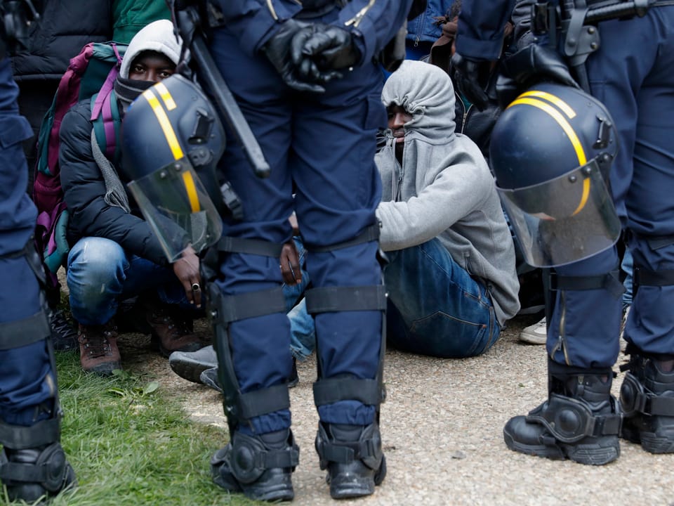 Polizisten stehen neben am Boden sitzenden Migranten