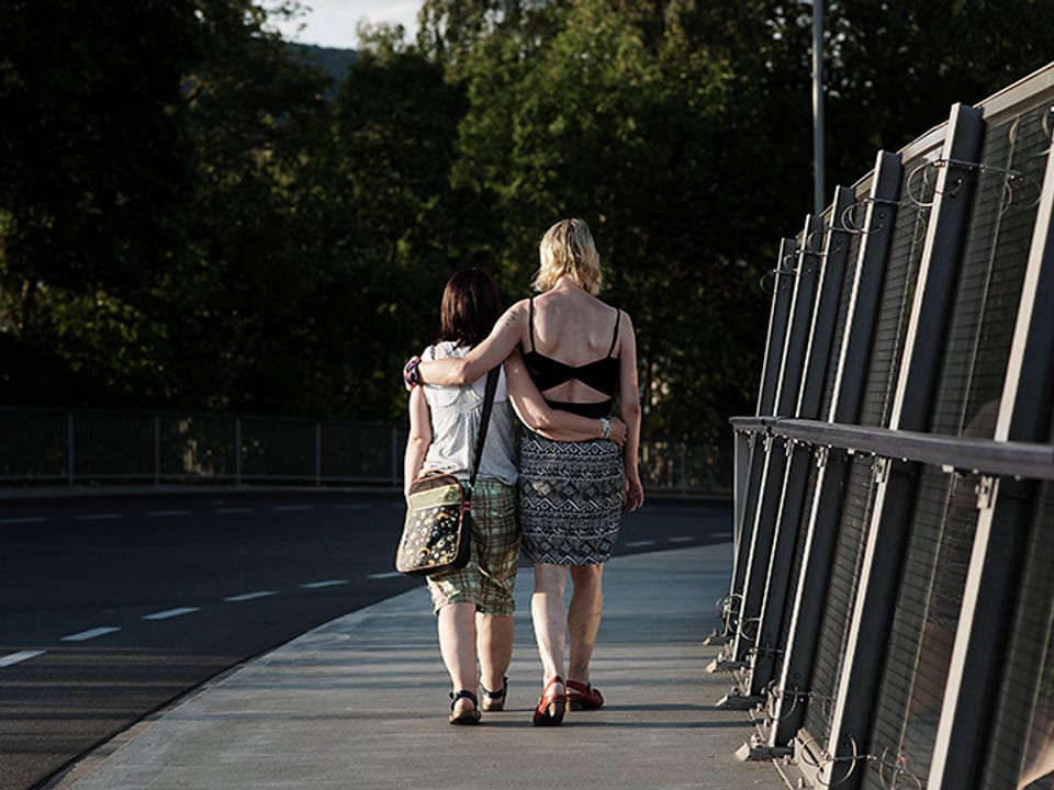 Zwei Frauen laufen Arm in Arm auf einem Trottoir, sie tragen sommerliche Kleidung.