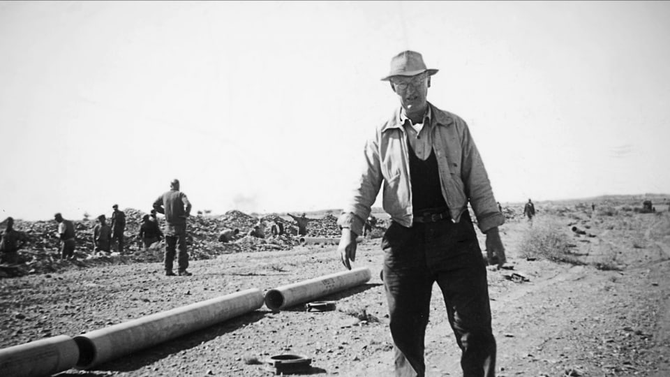 Ein altes Foto: Ein Mann steht in einer kargen Landschaft. Im Hintergrund arbeiten Menschen.
