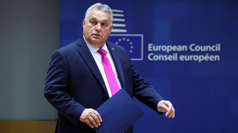 Viktor Orban trägt eine Mappe in der Hand und geht in einem Anzug am Logo der EU-Kommission vorbei.