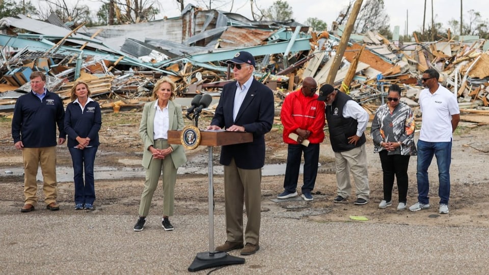 Mann steht in der Mitte an einem Rednerpult mit zwei Mikros. Dahinter sieben Personen und zerstörte Häuser.