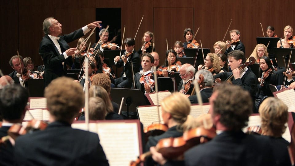 Ein Teil eines Orchesters, links im Bild der Dirigent, der zu jemandem zeigt