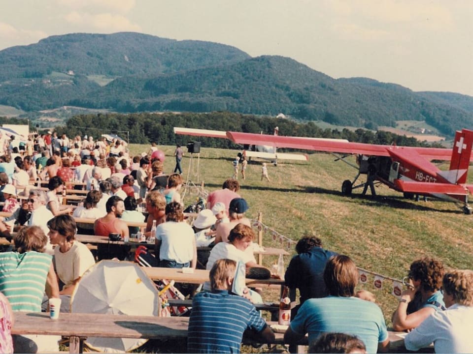 Altes Bild der Flugtage: Menschen sitzen an Festbänken, neben einem Flugzeug.