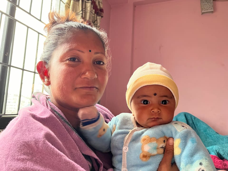 Eine Frau sitzt mit ihrem Baby in ihrem Arm in einem Raum. Beide blicken in die Kamera.